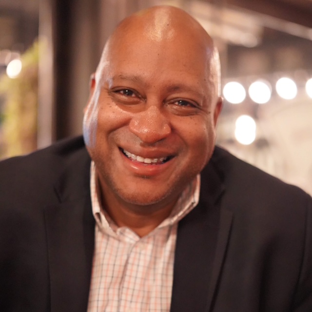Smiling man in a dark blazer and checkered shirt, looking at the camera in a warm indoor setting.